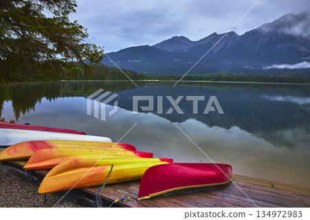 Colorful Canoes and Kayaks on Tranquil Lake with Mountain Reflections and Pine Trees Colorful Canoes and Kayaks on Tranquil Lake with Mountain Reflections and Pine Trees 134972983