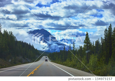 Highway Road Through Pine Forest With Dramatic Clouds And Snowy Mountain View 134973157