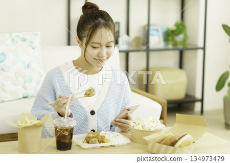 A young Japanese woman eating a bento lunch in the living room while looking at her smartphone and worrying about calories 134973479