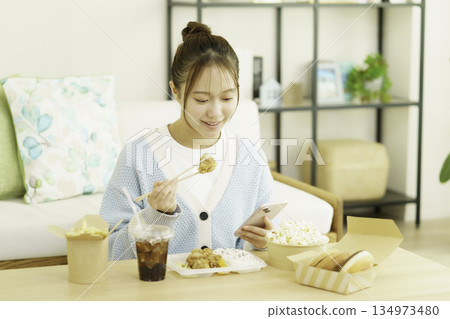 A young Japanese woman eating a bento lunch in the living room while looking at her smartphone and worrying about calories A young Japanese woman eating a bento lunch in the living room while looking at her smartphone and worrying about calories 134973480