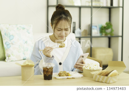 A young Japanese woman eating a bento lunch in the living room while looking at her smartphone and worrying about calories A young Japanese woman eating a bento lunch in the living room while looking at her smartphone and worrying about calories 134973483