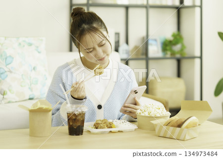 A young Japanese woman eating a bento lunch in the living room while looking at her smartphone and worrying about calories 134973484