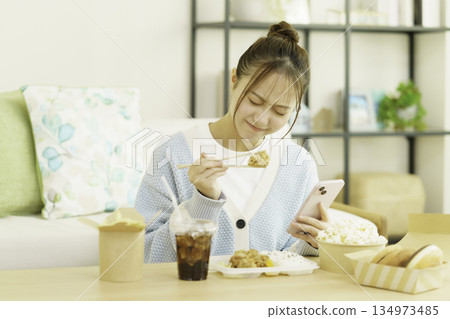 A young Japanese woman eating a bento lunch in the living room while looking at her smartphone and worrying about calories 134973485
