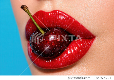 Close Up View Of Red Glossy Lips Holding A Fresh Ripe Cherry With Water Droplets On Blue Background 134973795