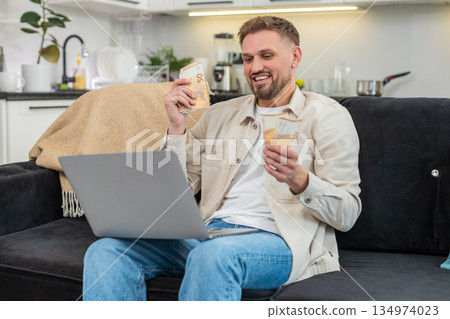 Young man counts cash euros compares with laptop earnings sheet fills sums smiles proud at goal met 134974023
