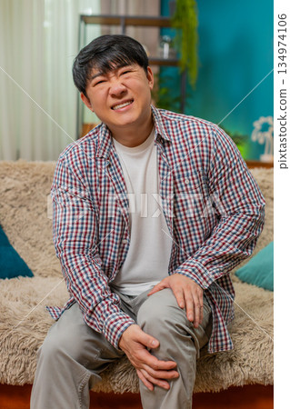 Asian man gripping knee with painful expression, massaging joint discomfort during movement struggle 134974106