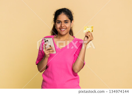 Indian woman holding credit card, focused on smartphone screen, making thoughtful online purchase 134974144