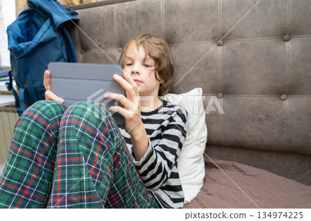 Boy in pajamas using a digital tablet in bed. Child focused on screen for e-learning Boy in pajamas using a digital tablet in bed. Child focused on screen for e-learning 134974225