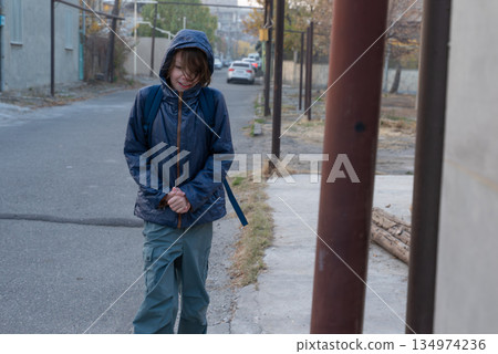 A boy in a hooded jacket shivers from the cold on an urban street. Student with backpack 134974236
