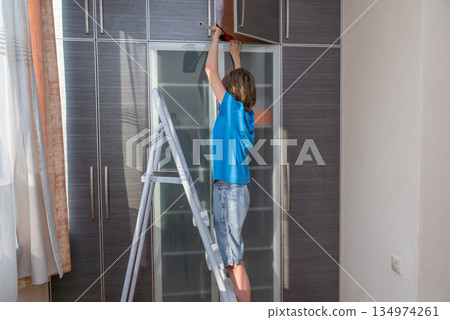 Boy helping with chores on a stepladder. Child cleaning the top of a modern wardrobe Boy helping with chores on a stepladder. Child cleaning the top of a modern wardrobe 134974261