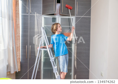 Young boy on a stepladder cleaning a wardrobe with a broom. Child helping with chores Young boy on a stepladder cleaning a wardrobe with a broom. Child helping with chores 134974262