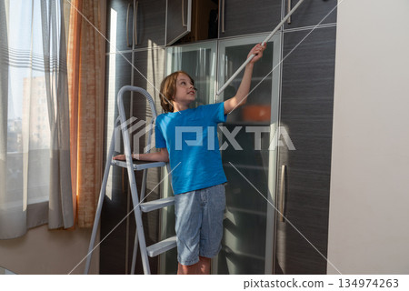 Young boy on a stepladder cleaning the top of a wardrobe. Child helping with household chores 134974263