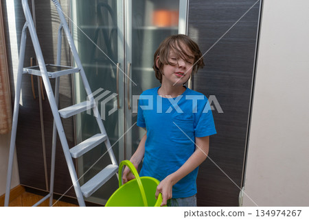 Tired boy with a bucket doing household chores. Unhappy kid helping with cleaning at home 134974267