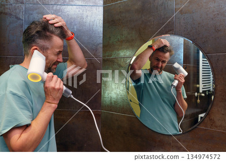 A man using a hairdryer in the bathroom. Daily grooming and self-care routine with mirror reflection A man using a hairdryer in the bathroom. Daily grooming and self-care routine with mirror reflection 134974572