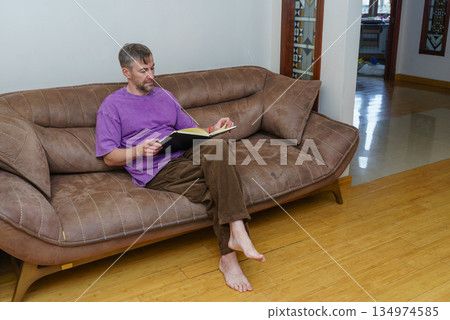 Middle-aged man reading a book while relaxing on a comfortable sofa in his living room Middle-aged man reading a book while relaxing on a comfortable sofa in his living room 134974585