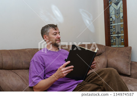 Casual man sitting on a sofa at home reading a book. Focused on learning and leisure Casual man sitting on a sofa at home reading a book. Focused on learning and leisure 134974587