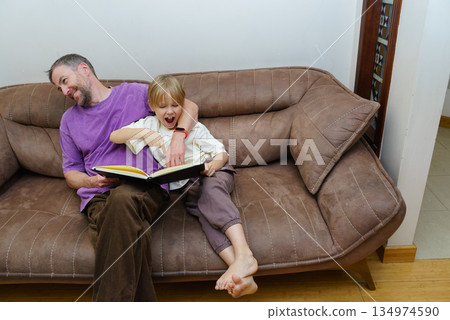 Excited young boy and his father reading a book together on a brown sofa. Happy family bonding 134974590