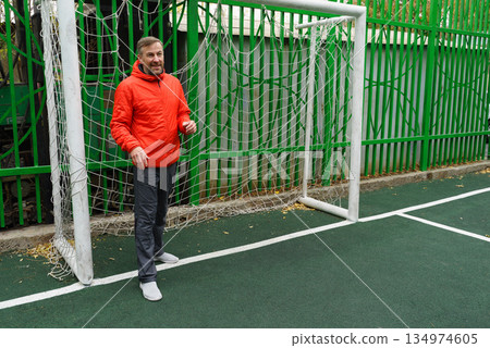 A smiling middle-aged man in an orange jacket stands in a soccer goal. Active lifestyle on an outdoor sports field. 134974605