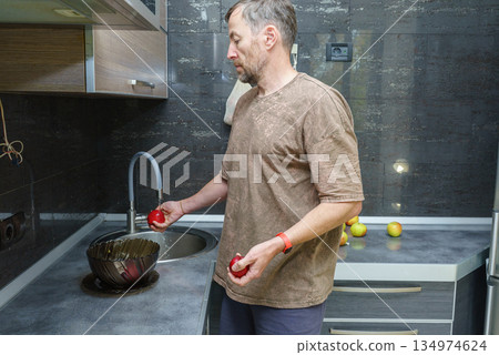Healthy food preparation. A middle-aged man washes fresh red apples in a modern kitchen sink 134974624