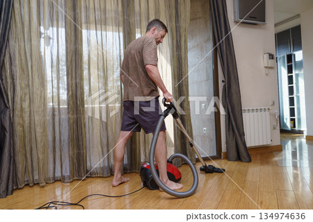 Man vacuuming the wooden floor at home. Domestic housework and cleaning concept 134974636
