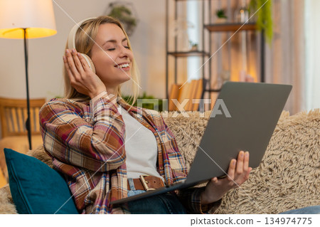 Smiling woman enjoying music on laptop at home with wireless headphones moving gently to rhythm Smiling woman enjoying music on laptop at home with wireless headphones moving gently to rhythm 134974775