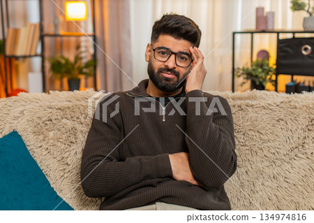Indian young man looks into camera with serious empathetic face on sofa at home, expressing warmth Indian young man looks into camera with serious empathetic face on sofa at home, expressing warmth 134974816