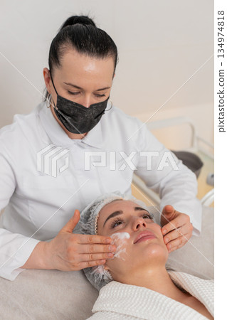 Cosmetologist applies cleansing foam to woman face during skincare massage procedure beauty clinic 134974818