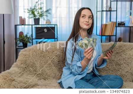 Young woman happily counting pile of cash dollar bills on sofa celebrating wealth successful finance Young woman happily counting pile of cash dollar bills on sofa celebrating wealth successful finance 134974823