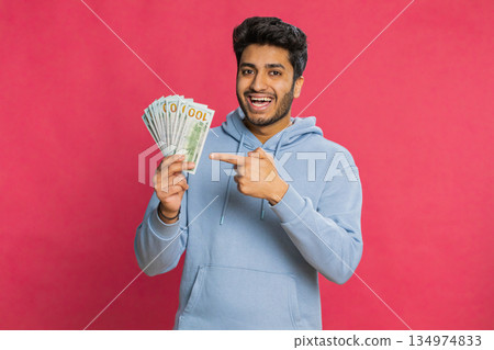 Rich Indian young man waving money dollar cash banknotes bills like a fan against red background Rich Indian young man waving money dollar cash banknotes bills like a fan against red background 134974833