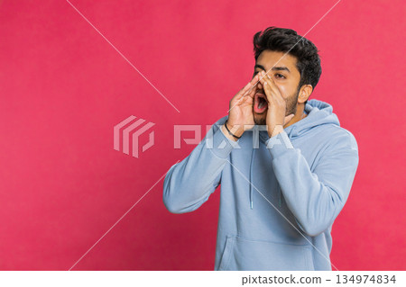 Indian young man shouting calling out loud holding hands near mouth yelling waving excited indoors Indian young man shouting calling out loud holding hands near mouth yelling waving excited indoors 134974834