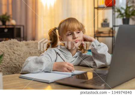 Tired young girl doing homework at table with laptop writing in notebook and showing lazy expression 134974840