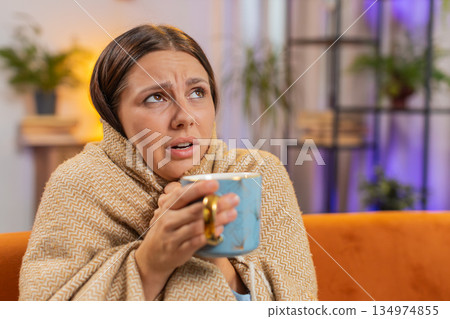 Young unhealthy sick ill woman covered by warm blanket drinks coffee freezes from cold flu at home Young unhealthy sick ill woman covered by warm blanket drinks coffee freezes from cold flu at home 134974855