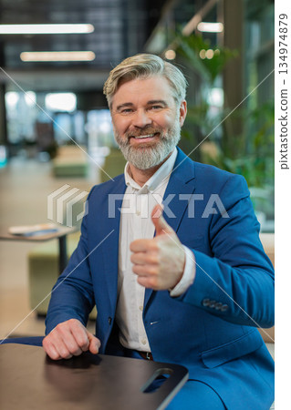 Happy mature Caucasian businessman in suit looking at camera showing thumb up in modern office lobby Happy mature Caucasian businessman in suit looking at camera showing thumb up in modern office lobby 134974879
