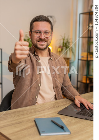 Happy man using laptop looking approvingly at camera showing thumbs up sitting at home office table 134974884