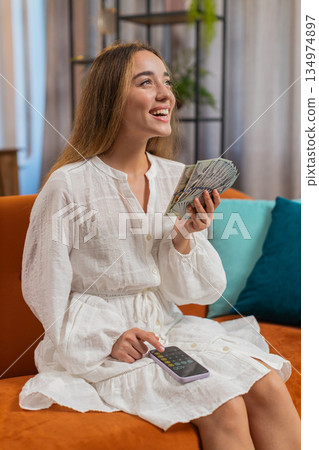 Rich happy young woman counting money cash using smartphone calculator sitting on sofa at home Rich happy young woman counting money cash using smartphone calculator sitting on sofa at home 134974897
