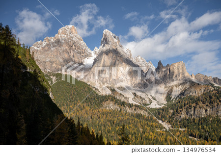 Dramatic rocky Dolomites mountain peaks above autumn forest under blue sky Dramatic rocky Dolomites mountain peaks above autumn forest under blue sky 134976534