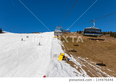 Austrian Alps ski resort shows strong contrast between white piste and snowless grass slope under blue sky. Climate warming, snow scarcity, chairlift infrastructure highlight winter tourism crisis 134976848