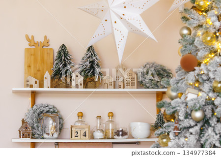 Christmas shelf decor with wooden houses, snowy fir trees and festive lights in kitchen interior 134977384