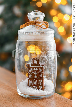 Christmas ornament gingerbread house in glass jar with snow on glowing lights illuminated background Christmas ornament gingerbread house in glass jar with snow on glowing lights illuminated background 134977390