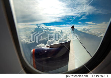 Scenic view of airplane wing and engine through passenger window flying above clouds under blue sky, concept of air travel vacation and transportation Scenic view of airplane wing and engine through passenger window flying above clouds under blue sky, concept of air travel vacation and transportation 134978001