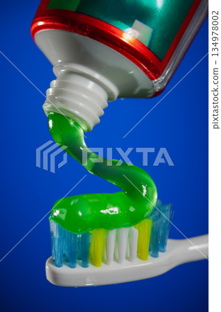 Green gel toothpaste being squeezed from tube onto colorful toothbrush bristles, dental hygiene and oral care concept on bright blue background. Green gel toothpaste being squeezed from tube onto colorful toothbrush bristles, dental hygiene and oral care concept on bright blue background. 134978002