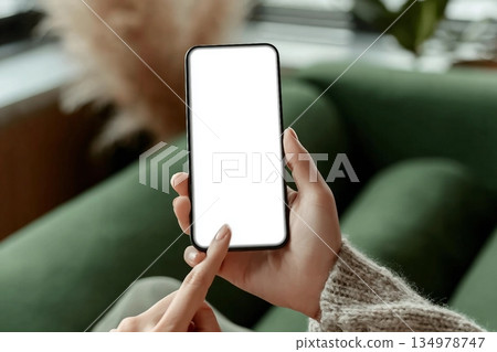 Close up of woman hand pointing on blank screen smartphone sitting on green sofa in cafe or lounge. Advertising Mockup, Social Media, Modern tech. Close up of woman hand pointing on blank screen smartphone sitting on green sofa in cafe or lounge. Advertising Mockup, Social Media, Modern tech. 134978747