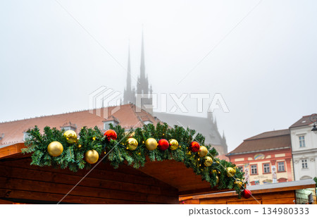 Decorated roof of a christmas market stall with fir branches and red and gold ornaments, brno city atmosphere with foggy silhouette of peter and paul cathedral in background Decorated roof of a christmas market stall with fir branches and red and gold ornaments, brno city atmosphere with foggy silhouette of peter and paul cathedral in background 134980333