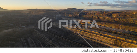 Ultra high resolution panoramic view of an open pit coal mine with massive mining machines, layered terrain, and dramatic sky on the horizon. Powerful industrial landscape showing large scale Ultra high resolution panoramic view of an open pit coal mine with massive mining machines, layered terrain, and dramatic sky on the horizon. Powerful industrial landscape showing large scale 134980452