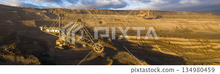 Ultra high resolution panoramic view of an open pit coal mine with massive mining machines, layered terrain, and dramatic sky on the horizon. Powerful industrial landscape showing large scale 134980459