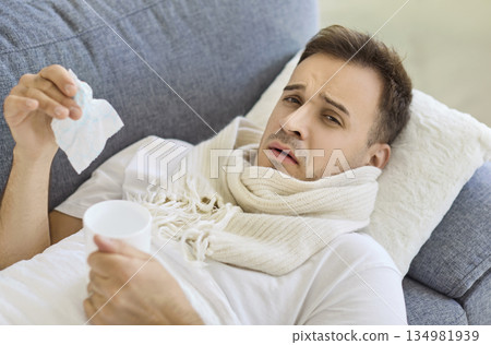 Sick man with scarf holding tissue and cup, resting on sofa at home interior Sick man with scarf holding tissue and cup, resting on sofa at home interior 134981939
