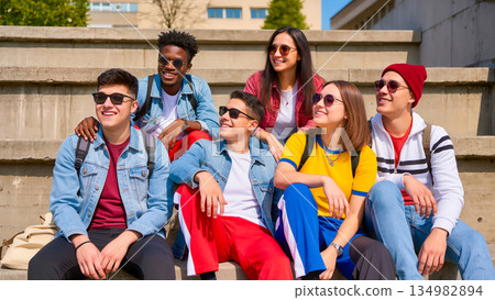 Six young people (boys and girls) of different nationalities sit on concrete steps on a sunny day. The students are dressed in bright casual clothes, many wearing sunglasses. They smile and look in th 134982894
