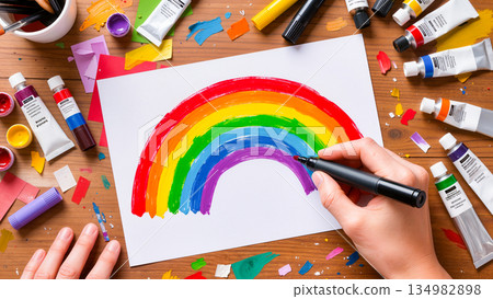 An overhead view of the creative process. Paint tubes, scraps of colored paper, and brushes are laid out on a wooden table. In the center is a white sheet of paper with a seven-color rainbow drawn on  134982898