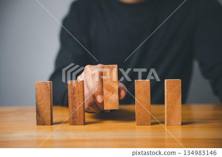 A hand places a wooden block in a row of four on a table. The blocks are made of natural wood and have a grain pattern. The table is made of a light wood. . 134983146