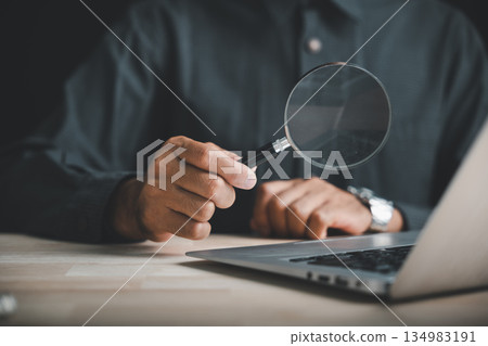 Closeup of a hand holding a magnifying glass over a laptop keyboard, searching for information online. 134983191